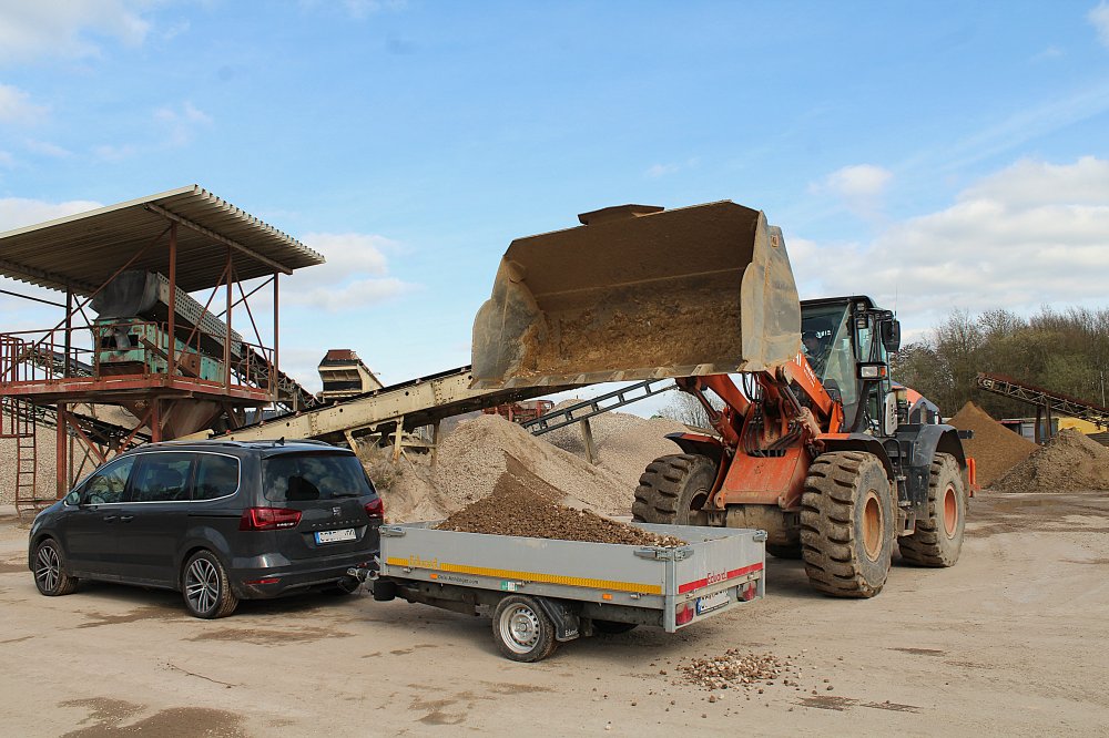 Verladen von Kleinmengen, Selbstabholung Ein Pkw-Anhänger wird per Radlader/Bagger mit Schotter beladen